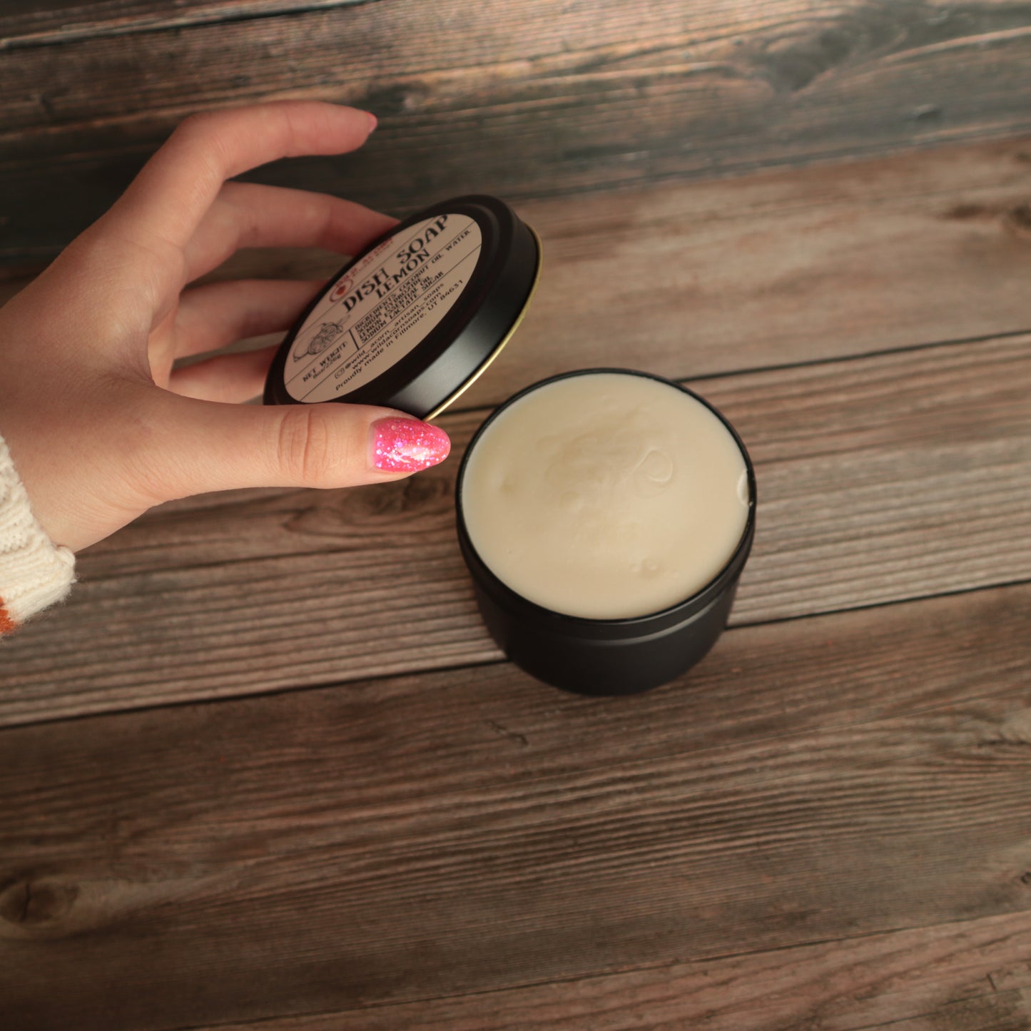 black candle like jar with lid open holding a pale yellow solid dish soap, lid being held open by a hand