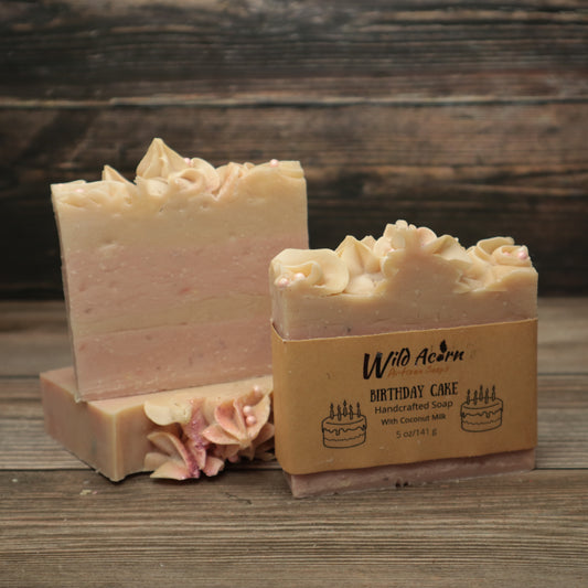Three bars of soap with pink and white layers, frosting appearance on top with biodegradable glitter and pink pearl sugar sprinkles. One bar has a label on it with pictures of birthday cake with candles on it.