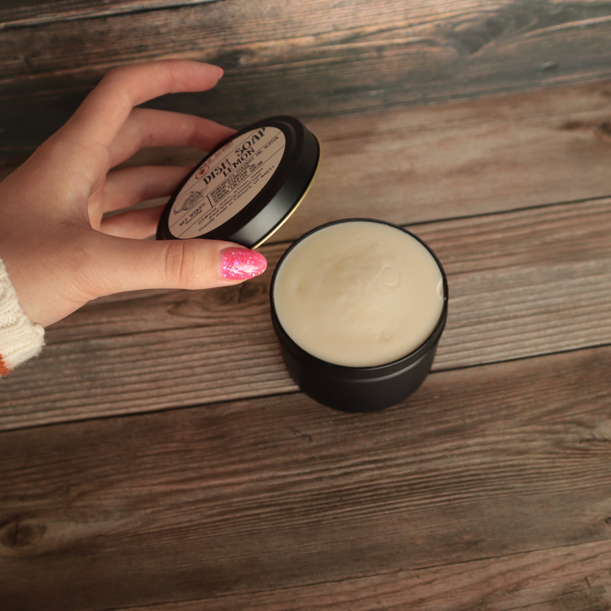 black candle like jar with lid open holding a pale yellow solid dish soap, lid being held open by a hand